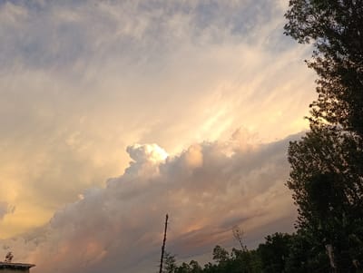 The image shows a cloudy sky with a vibrant pink and yellow sunset in the background. The sun's rays shine through the clouds, creating a bright, ethereal effect. Trees and foliage are visible in the foreground, with a tall, thin post standing in the middle ground. The image was taken in Kaplelwo, Kenya, at the coordinates -0.416, 35.146.