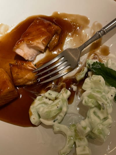 A partially eaten meal is on a white plate, featuring glazed salmon and cucumber salad. Brown sauce pools around the salmon. A silver fork rests on the plate, piercing a piece of salmon. Creamy, white dressing coats the diced cucumbers, speckled with black pepper or herbs. A single green leaf garnish sits atop the salad.