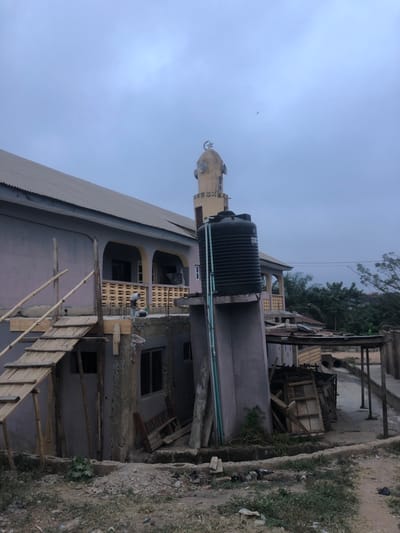 A mosque's minaret pierces a cloudy sky. A large, black water tank sits atop the building, connected by green pipes. The structure appears to be under construction, with scaffolding and wooden planks visible. A pile of debris and building materials clutters the ground nearby. Trees and vegetation surround the mosque, creating a contrast between the natural and built environment.