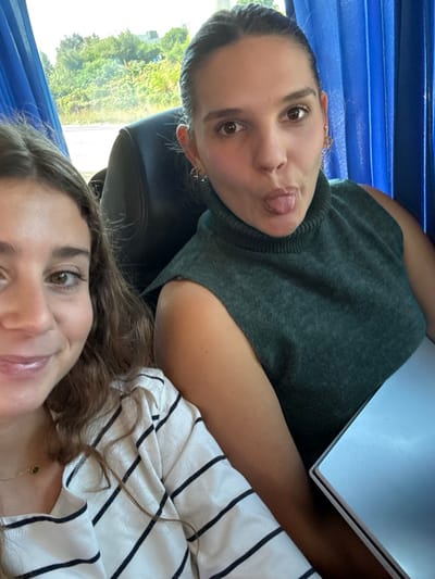 Two females are depicted inside a vehicle, likely a bus, located in Amadora, Portugal. The interior features blue fabric window coverings and dark upholstered seating. Through a window on the left, a blurred view of green foliage and an indistinct structure or road is visible, indicating a daytime setting.

In the foreground, positioned to the left, a female subject is partially visible, showing her lower face, neck, and torso. She has light brown hair and a slight smile. She wears a white long-sleeved shirt with horizontal black stripes and a thin gold-colored necklace featuring a small green circular pendant.

Behind and to the right, a second female subject is visible from the chest up. She has light brown hair pulled back from her face. Her ears display multiple small hoop earrings. She wears a dark green sleeveless turtleneck top. Her expression is characterized by a playful gesture, with her tongue protruding from her mouth, and she is looking directly towards the camera. A silver-colored laptop is partially visible on the bottom right, positioned on a surface or lap in front of her.