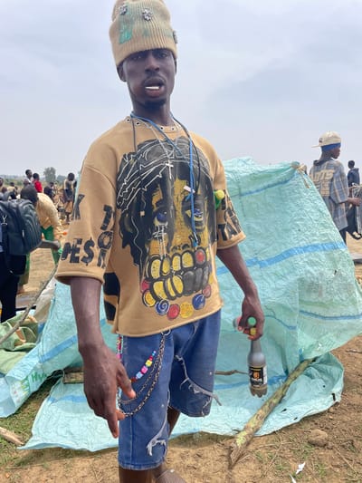 A man stands in a field, holding a bottle of "Fearless" drink in his left hand. He is wearing a decorated knit cap, a tan long-sleeved shirt with an image of a lion and the word "Fearless" on the sleeve, and blue denim shorts. A silver chain with colorful beads is attached to his shorts. Behind him is a pile of material covered with a light blue tarp with blue stripes. In the background, other people are visible. The location is Maiyanga, Nigeria. The ground is dirt and grass.