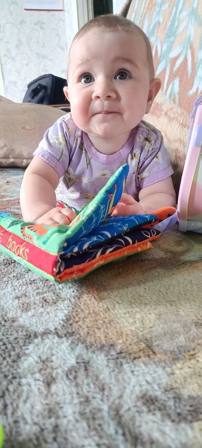 A baby sits on a patterned rug, facing forward with their head tilted upwards and to the left. The baby is wearing a light purple t-shirt with a floral pattern and has wide, dark eyes, a small nose, and slightly pursed lips. Their hands are resting on a folded fabric book with colorful patterns and red trim, on which the word "books" is partially visible in red lettering. Behind the baby, a light-colored couch with a textured pattern is visible. To the left of the baby, a black bag is partially visible. To the right, a pink and blue object, possibly a toy or a stroller part, is present. The scene appears to be indoors, in what could be a living room or play area, with a textured rug covering the floor and a patterned fabric in the background. The location is described as Каменное, Russia.