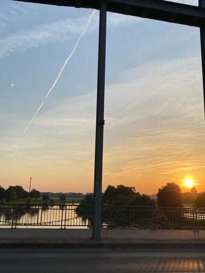 The image depicts a sunset scene viewed from a bridge in Rinteln, Germany. The sun is low in the sky, casting a warm orange and yellow glow across the clouds and the water below. A contrail from an airplane stretches across the sky.
The foreground features a dark metal structure, likely part of the bridge's framework, and a railing with numerous locks attached, a common sight on bridges known for romantic traditions. Beyond the railing, a calm body of water reflects the sunset colors. The opposite bank of the river is lined with trees and greenery, with a few buildings and a telecommunications tower visible in the distance. The overall mood is serene and peaceful, enhanced by the soft light of the setting sun. The time of day is clearly sunset. The weather appears to be clear with some wispy clouds. There is no visible text in the image.