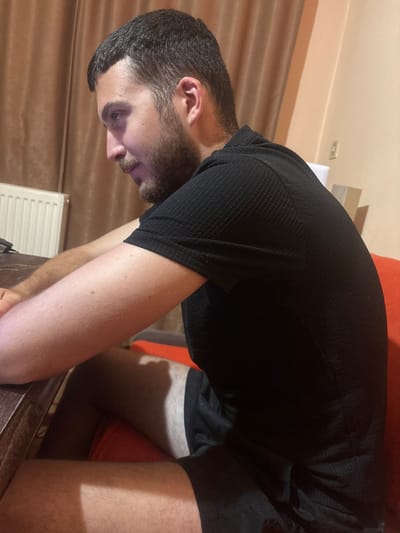 A man is seated indoors, viewed from the side in a medium shot. He has short dark hair, a beard, and is wearing a black t-shirt and black shorts. He is leaning forward, with his arms resting on a wooden table. A radiator is visible behind him, along with brown curtains and a beige wall. The scene appears to be a residential interior in Каменар, Bulgaria.