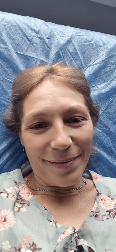 A medium-shot, close-up selfie shows a woman with light brown hair and fair skin, lying down on a blue, crinkled fabric. The woman is looking directly at the camera with a slight smile. She is wearing a light blue, floral-print top with a silver chain necklace. The background is dominated by the blue fabric, which appears to be a disposable sheet or gown. The lighting is even and appears to be artificial, casting no harsh shadows. The overall impression is that of a casual, personal photograph taken in a private setting. There is no indication of the specific location within the city of Екпінді, Kazakhstan.