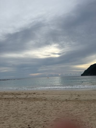 This is a photograph of a beach in Meunasahbale, Indonesia, taken during overcast conditions. The foreground is dominated by sandy beach with some scattered debris. The middle ground shows the gentle waves of the ocean meeting the shore, with some white foam visible. In the distance, the ocean stretches to the horizon, with a small yellow object, possibly a boat or buoy, visible on the water. To the right, a dark, tree-covered hill slopes down to the sea. The sky is filled with heavy, gray clouds, with a hint of lighter sky and possibly the sun peeking through on the horizon. There are a few people visible in the water, appearing to be swimming or surfing.