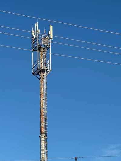 An outdoor, medium-shot, vertical, eye-level, slightly-low-angle, full-body photograph captures a cell tower against a clear, bright blue sky in Gavrilov Posad, Russia. The cell tower, a tall, slender, metal structure, is positioned off-center to the left of the frame. It is comprised of a white, lattice-like frame extending upwards. Several white, rectangular antennas are mounted on the upper section of the tower. A vertical ladder with white rungs runs along the length of the tower. The tower's central pole exhibits significant rust and discoloration, indicating age or exposure to the elements. Four parallel, thin, metallic wires run horizontally across the upper portion of the frame, passing in front of the cell tower and the sky. A portion of another wire structure with small, dark electrical components is visible in the lower right corner of the image. The scene is devoid of any people or animals.