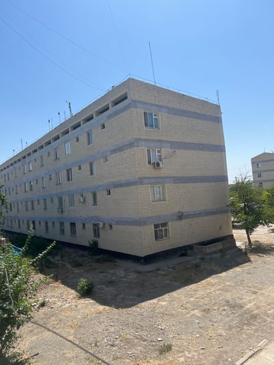 This image captures a view of a large, multi-story apartment building constructed with light-colored bricks and horizontal grey bands. The building has a flat roof with several antennas and a railing. The windows on the building are rectangular, and some have air conditioning units mounted outside. In the foreground, there is a dry, dusty ground with sparse vegetation and rocks. The background shows a clear blue sky and the upper portion of another similar apartment building and some trees. The scene appears to be in an urban or semi-urban environment, likely residential. The location is identified as Şamaldı-Say, Kyrgyzstan.