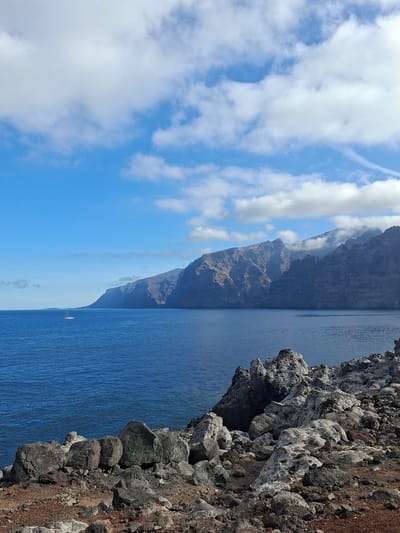 The image shows a scenic coastal view in Puerto de Santiago, Spain. In the foreground, there is a rocky, uneven terrain with various sized stones, ranging in color from dark gray to reddish-brown. Beyond this rocky shore, a vast expanse of deep blue ocean stretches towards the horizon. A small white sailboat is visible in the distance on the water. Along the far edge of the ocean, a dramatic mountain range with steep cliffs rises sharply, partially shrouded in clouds at its peaks. The sky above is a clear, bright blue, dotted with fluffy white clouds, some of which are thin and wispy, while others are more substantial. The overall scene depicts a rugged and natural coastal landscape under a bright sky.
