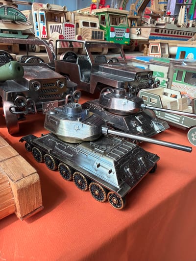 This image captures a close-up view of several metal model tanks displayed on an orange fabric surface, likely in a market stall or shop in Jiejinkou, China. The perspective is from above and slightly to the side, with the image rotated 90 degrees counter-clockwise from its natural orientation.

In the foreground, three intricately detailed metallic models, resembling military tanks or vehicles, are arranged. They are primarily dark gray or bronzed metal, with some lighter, shinier silver-toned components, particularly the turrets and gun barrels. Each tank features individual track wheels with radial designs, textured body panels, and protruding gun barrels. One of the tank turrets distinctly displays a star emblem. These models appear to be sturdy, possibly made from cast metal or heavy alloys, and show slight signs of wear.

Behind these metal models, the background is filled with a dense array of wooden models or toys. These items are crafted from light-colored wood and painted with various bright colors, including green, blue, red, and orange. They depict miniature buildings, vehicles, and possibly train components, suggesting a collection of educational toys or collectibles. Details such as windows, doors, and patterned designs are visible on these wooden objects. One of the wooden structures appears to have the partial word "TURBO" visible.

The entire arrangement rests on a textured, plain orange fabric, which could be a tablecloth or display mat. Natural or artificial light illuminates the scene, casting soft shadows on the fabric, particularly around the tank models. A rough-hewn wooden block or stand is partially visible in the bottom right corner. There is no discernible activity taking place, rather the objects are static, presented for viewing or sale. No people are visible.