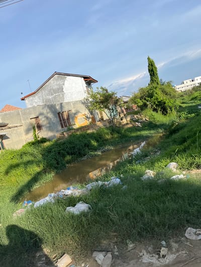 This is a photograph taken outdoors on a sunny day in Landuh, Indonesia. The image shows a small, muddy stream running through a grassy area. There are several houses visible in the background, some of which appear to be in disrepair. There is also a significant amount of trash and debris scattered along the banks of the stream and in the surrounding vegetation. The sky is blue with some scattered clouds. A tall, slender tree stands prominently in the middle ground. The overall impression is one of a somewhat neglected urban or semi-urban environment.