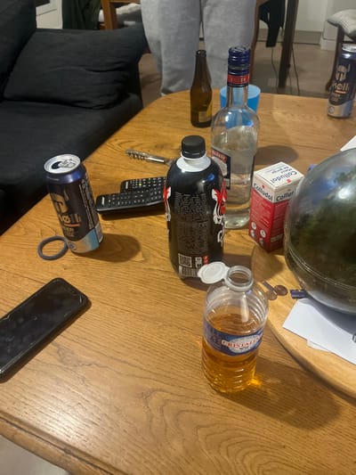 The image displays a wooden table laden with various items in what appears to be a domestic living space in Amiens, France. A person wearing grey sweatpants is partially visible standing in the background, situated near a dark grey sofa.
On the table, from left to right, are: a dark-colored smartphone with a black screen, a dark elastic hair tie, an open blue and silver Mikkeller beer can, two black remote controls, a silver corkscrew, a large black bottle with a white and red tiger graphic (likely an energy drink), a clear glass bottle of Poliakov vodka that is partially filled, and a red and white box labeled "Colludol Maux de Gorge" (sore throat medication). Further right, an open Cristaline water bottle contains an amber-colored liquid, with two copper coins resting nearby on a white paper or napkin. A large clear glass spherical terrarium with green plant material inside is also present. In the background, another Mikkeller beer can, an empty brown glass bottle, and a small blue plastic cup are visible. The arrangement suggests a casual gathering.