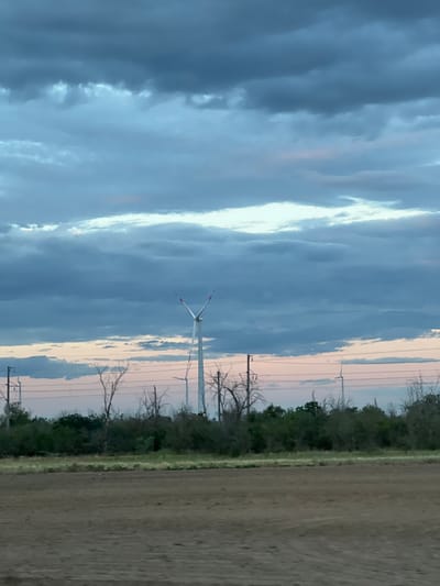 The image depicts a landscape scene with a cloudy sky and wind turbines. The sky is filled with dramatic, dark blue and grey clouds, with a bright white streak of light cutting through them. In the distance, several wind turbines stand tall, silhouetted against the lighter parts of the sky. The turbines have white blades with red tips, and one is prominently visible in the center of the frame.
The foreground on the right side of the image shows a section of a field, appearing to be agricultural land, with a line of trees and vegetation separating it from the sky. The trees are bare of leaves, suggesting it might be autumn or winter. There are also power lines and poles visible, indicating human infrastructure in the area.
The overall impression is one of a vast, open landscape under a dynamic sky. The lighting suggests it is either early morning or late evening, given the soft, diffused light and the colorful hues in the sky, particularly where the clouds meet the horizon, showing hints of pink and peach. The weather appears to be changing or stormy, judging by the heavy clouds. The scene evokes a sense of industrial power set against a natural, somewhat austere environment. There is no visible text in the image.