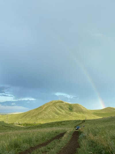 The image depicts a vast, rolling green landscape under a partly cloudy sky with a faint double rainbow visible. A dirt track winds its way through the tall grass, leading towards a prominent, rounded green hill in the middle ground. Atop this hill, a solitary white tower stands. Further along the track, a dark blue SUV is visible, its headlights on, with two people standing near it. The environment suggests a rural or natural setting, possibly on the outskirts of Верхнеозёрное, Russia, given the presence of the tower, which could be a telecommunications or observation tower. The overall mood is serene and tranquil, with the natural beauty of the landscape dominating the scene.