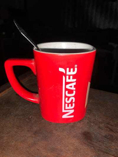 A close-up, low-light, eye-level shot shows a red Nescafe mug with a spoon resting inside it, sitting on a dark, wooden surface. The mug is partially filled with a dark liquid. The Nescafe logo, in white, is visible on the side of the mug. The background is dark and out of focus. The image does not depict the city of Martove, Ukraine, or any specific urban environment.