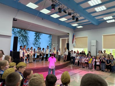 In an indoor assembly hall in Pron'kino, Russia, a group of children and young adults are gathered on and around a stage. A large projection screen displays an image of snow-covered trees. On the wooden stage, a group of boys and girls, some wearing blue scarves, stand in a line facing the audience. A young woman in a pink shirt and jeans stands in front of them, facing away from the camera, appearing to address the group. To the right of the stage, several adults and children are also present, some holding a Russian flag. The hall is brightly lit by fluorescent ceiling lights and stage spotlights, casting pink and white hues on the floor. The environment suggests a school or community event.
