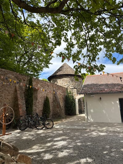 The image depicts a view in Častá, Slovakia. A stone building with a rounded tower stands decorated with colorful bunting. Bicycles are parked against the wall of this building. A tree with dense green foliage occupies the left side of the frame. A path leads beside the building, and a white structure is visible to the right, along with a gravelly area. An old wooden wheel leans against the wall. The sky shows patches of blue between the foliage.