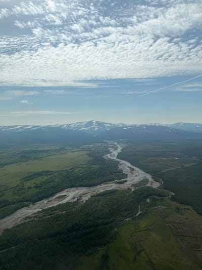 An aerial view of a vast, sparsely populated landscape with a wide, meandering river flowing through it. The riverbed appears to be sandy or gravelly, with multiple braided channels. Lush green forests and open fields surround the river, transitioning into a mountainous region in the distance, capped with snow. The sky is partly cloudy with scattered white clouds and a clear blue sky. The scene depicts a remote natural environment, likely in the wilderness of Stekolny, Russia, with no signs of human activity other than a faint contrail in the sky.