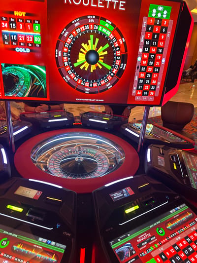 Here is a description of the image:
**Location:** The image appears to be taken in a casino, likely at the Seminole Hard Rock Hotel & Casino on the Hollywood Seminole Reservation, Florida, USA.
**Subjects and Objects:** The primary focus is on several automated roulette machines. These machines feature digital displays showing the roulette wheel, betting options, game statistics, and "hot" and "cold" numbers. The physical roulette wheels themselves are visible within the circular machines. A chair is partially visible in the background. The reflections in the glass surfaces of the machines suggest a brightly lit environment and possibly other casino patrons nearby.
**Details:**
* The digital displays of the roulette machines show various betting options and odds.
* The "hot" and "cold" number indicators display which numbers have recently appeared more or less frequently.
* The roulette wheels are in motion, with the ball visible in some of the wheels.
**Environment:** The casino environment is evident from the design of the machines, the lighting, and the overall ambiance suggested by the reflective surfaces. The details suggest a high-end casino setting.
