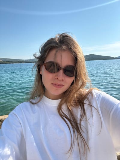 A young woman with long, light brown hair and sunglasses takes a selfie outdoors with a body of water and distant islands in the background. She is wearing a white t-shirt. The water is a clear turquoise color and appears to be slightly wavy. The sky is bright blue with some sun glare visible. The scene suggests a sunny day, possibly at a coast or lake. While the image does not explicitly depict the city of Grad Šibenik, Croatia, such coastal scenery is common in that region.
