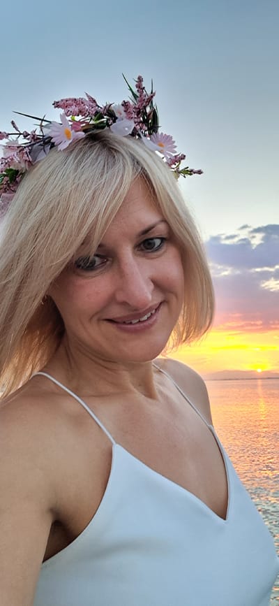 A woman with blonde hair and a flower crown is taking a selfie at sunset on the coast of Agia Triada, Greece. She is wearing a white spaghetti strap dress. The sky is filled with colorful clouds, and the sun is setting over the ocean. The water is calm, and there is a distant shoreline visible.