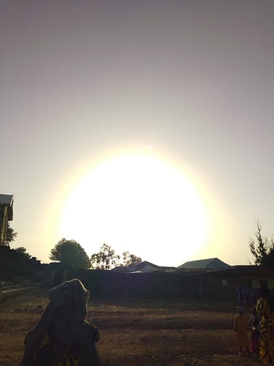 An outdoor scene in Bahit, Nigeria, captured under bright, central sunlight creating a prominent halo effect in the sky. The foreground features a large, dark, silhouetted figure in the lower left, wearing a head covering and facing away from the camera. The ground is dry, reddish-brown earth. In the midground to the right, several smaller figures, including what appear to be children in colored clothing (yellow, pink, patterned), are partially visible. A low, dark wall or structure extends horizontally across the midground. Behind this wall, multiple single-story buildings with varied rooflines are discernible, partially obscured by tree foliage. Additional trees are visible as silhouettes against the bright sky on the left and right sides of the frame.