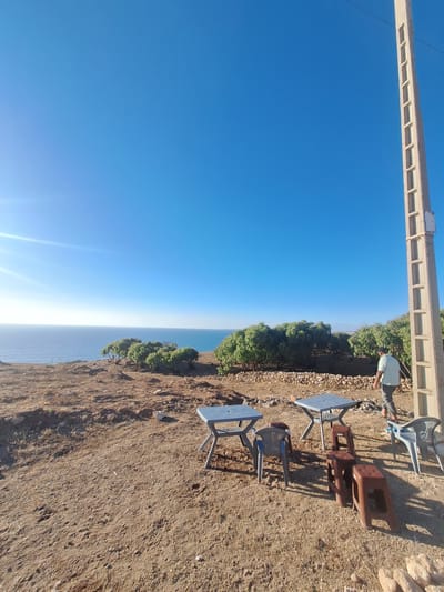 This is a sideways, outdoor photograph of a scenic overlook near Ahrara, Morocco. The scene is dominated by a bright blue sky and the calm, blue sea stretching to the horizon. In the foreground, a person in a light blue shirt and dark pants stands near a low, rocky wall, looking down towards the sea. Scattered on the dry, earthy ground are several outdoor tables and chairs, some made of metal and plastic, others of brown plastic. One table has a blue top, and another appears to be made of dark, possibly wood, material. The ground is covered in dry soil, small stones, and sparse vegetation, suggesting a warm, arid climate.

A concrete structure, possibly a bridge or elevated walkway with rectangular openings, runs horizontally across the top of the frame. Trees with lush green foliage grow along the edge of the cliff, providing a stark contrast to the dry ground. The sun is high in the sky, casting distinct shadows, indicating it is likely midday. The overall mood is peaceful and serene, with the vastness of the sea and sky creating a sense of tranquility. There is no visible text in the image.