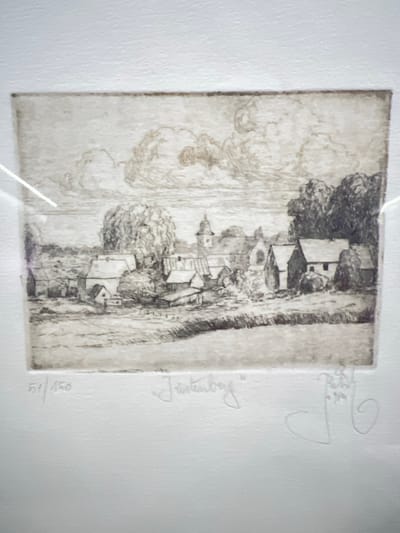 The image is an etching depicting a village scene. In the foreground, a field stretches towards a cluster of buildings, likely houses and a church with a steeple. Trees are scattered throughout the landscape, with a particularly large, bushy tree on the left and a denser grove on the right. The sky is filled with dramatic, swirling clouds.

The etching is signed in the lower right corner with a stylized signature and dated "1964". To the left of the signature, the etching is numbered "51/150". The overall style is detailed and atmospheric, suggesting a rural setting. The location in the city of Schoonderbuken, Belgium, is not evident from the image itself, and this information would need to be provided externally.