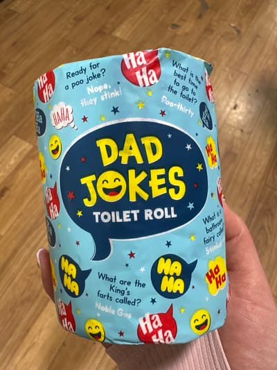 A roll of novelty toilet paper, wrapped in a light blue paper with a repeating pattern of "Ha Ha" speech bubbles, yellow smiley faces, and red and yellow stars, is held by a person's hand. The main text on the roll reads "DAD JOKES TOILET ROLL" in large yellow letters against a dark blue speech bubble. The paper also features several jokes, including "Ready for a poo joke? Nope, they stink!", "What is the best time to go to the toilet? Poo-thirty", and "What are the King's farts called? Noble Gas". The background is a wooden floor. This image does not provide any information about the location in the city of Carlow, Ireland.