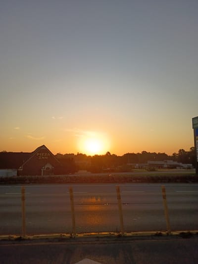 The image depicts a sunset over a road, likely in Frankfort, United States of America. The sun is a bright, yellow orb low on the horizon, casting a warm, orange glow across the sky and illuminating the surrounding trees and buildings. The sky transitions from a pale blue at the top to a vibrant orange and yellow near the sun.
In the foreground, a road stretches horizontally across the frame, with visible lane markings. The road surface appears wet and reflective, suggesting recent rain or dew. Along the side of the road, yellow posts with reflective tape are placed at intervals, likely serving as traffic markers or delineators. A Pizza Inn restaurant building is visible to the left of the road, its roofline sharply defined against the sunset. The building is constructed of brick and has a sign with "PIZZA INN" written vertically on its gable end. Further back, a line of trees and some buildings are silhouetted against the colorful sky.
The overall scene conveys a sense of calm and tranquility, typical of a sunset. The light suggests it is either early morning or late evening. No people are visible in the frame. The visible text includes "PIZZA INN" on the restaurant's sign and partial text on a distant sign that reads "BRIGGS SHOP" and "SHOPPING".