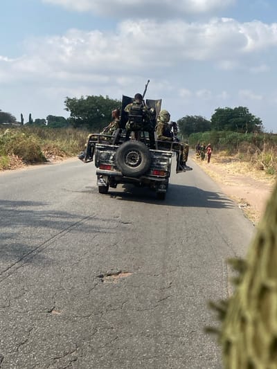 The image depicts a security situation on a partially degraded asphalt road in Sankara, Nigeria, under a partly cloudy sky. In the midground, a dark-colored Toyota pickup truck, modified into a technical, moves away from the viewer. Mounted in the truck bed is a heavy machine gun, operated by a standing individual in camouflage uniform and body armor. Two other armed individuals in camouflage uniforms are seated on the sides of the truck bed; one on the right wears a light-colored face-covering headwrap. A spare tire is mounted on the truck's tailgate. The road shows cracks and potholes. Along the right roadside in the background, a group of approximately five to seven civilians, dressed in various colored clothing, walk in the same direction as the truck. The environment is characterized by dry, sparse vegetation and scattered trees bordering the road. The immediate foreground on the right side shows a blurry, textured object, possibly part of another camouflage uniform, suggesting the perspective of the observer.