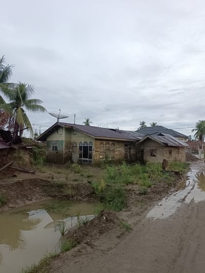 The image depicts a dilapidated scene in Meunasah Raya, Indonesia, characterized by overcast skies and a waterlogged environment. In the foreground, a muddy path with large puddles of water reflects the surroundings, including palm trees and a satellite dish. Adjacent to the path is a small, stagnant pond filled with murky water and scattered aquatic vegetation.

Dominating the mid-ground are two distressed structures. The larger building, appearing to be a house, has a faded, greenish-blue exterior that is peeling and stained. Its roof is in disrepair, with some sections missing or damaged. A satellite dish is prominently mounted on the roof. The windows are either boarded up or broken, and the overall structure shows signs of neglect and decay. To the right of this main house is a smaller, more rudimentary structure with a rusty metal roof and walls made of rough concrete or plaster. This building also appears abandoned and in poor condition.

The area surrounding the buildings is overgrown with weeds and sparse vegetation. Debris, including what appears to be broken metal sheeting and scattered wooden planks, is visible, particularly to the left of the larger house, suggesting past damage or demolition. In the background, other houses with tiled roofs are visible, some appearing more modern and intact than the foreground structures, hinting at a residential area. Palm trees are scattered throughout the scene, characteristic of the tropical climate. The overall impression is one of poverty and neglect, possibly exacerbated by recent flooding or a general lack of maintenance. No people are present in the image.