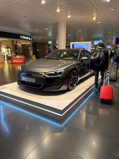 The image captures a scene at Munich Airport (MUC) in Oberding, Germany, where an Audi RS e-tron GT is on display. The car is positioned on a raised platform with integrated blue lighting. A man with a backpack is standing beside the car, looking at it. He has a red suitcase next to him. In the background is a "MyDutyFree" store. The floor is tiled and glossy. Other people and luggage are visible in the background.