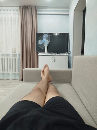 This is a first-person perspective, point-of-view shot taken from a couch in what appears to be a residential living room in Ürjar, Kazakhstan. The scene is brightly lit, likely by indoor lighting, and depicts a relaxed, casual atmosphere.

In the foreground, the subject's legs and feet are visible, resting on the couch. The subject is wearing black shorts, and their bare feet are crossed at the ankles. The couch itself is a light beige or cream color, upholstered in a textured fabric.

The middle ground of the image reveals a white cabinet with two doors, topped by a black flat-screen television. A white fan with blue blades stands on the cabinet to the left of the television. Behind the cabinet and television, the wall is painted a light blue.

To the left of the scene, sheer white curtains are drawn across a window, and behind them, heavy brown curtains are partially visible. A white radiator can be seen beneath the window. The flooring appears to be light-colored laminate or wood. The room seems tidy and well-maintained.