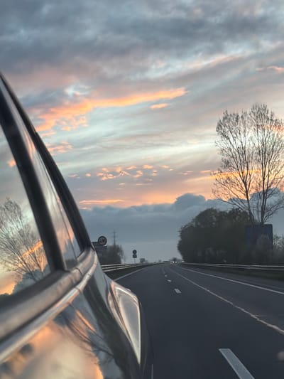 The image shows a view from inside a car, looking out the driver's side window. The car is traveling on a highway. The sky is filled with clouds illuminated by a sunset or sunrise, creating orange and pink hues. A bare tree is visible in the distance. Road markings are present on the asphalt, and a roadside sign is visible further ahead. The reflection of the sky and tree is visible in the car window. The location cannot be confirmed with the information given in the image.