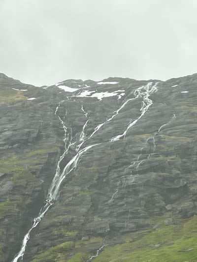 The media file depicts a close-up view of a sheer, grey rock face with patches of green vegetation clinging to its surface. White streaks, likely water, cascade down the rock face in a series of rivulets, creating a dynamic, textural contrast against the darker stone. The rock appears rugged and weathered, with visible striations and crevices. The upper left portion of the image is a bright, overcast sky, suggesting a cloudy or foggy day. The overall impression is one of a dramatic natural landscape, possibly a cliff face or mountainside in Norway, given the location context of Røldal. There are no people or man-made objects visible. The lighting is diffused, consistent with an overcast sky, and there is no indication of a specific time of day. The mood is serene and majestic, emphasizing the raw beauty of nature. No text is visible in the image.