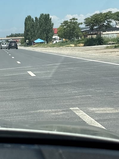 The image depicts a road scene in Shahumian, Armenia, viewed from inside a vehicle. The road is paved and has white lane markings. On the left side of the road, there are trees, buildings with red roofs, and solar panels. A blue tarp is visible amongst the trees. Several cars are parked or driving on the side of the road. The sky is blue with some clouds, suggesting it is daytime with clear weather. The overall scene appears to be a rural or semi-rural area. There is no visible text in the image.