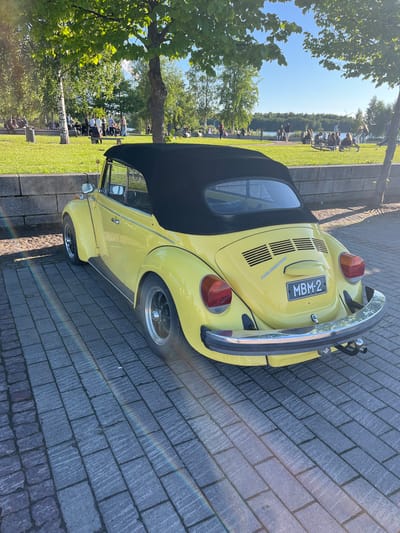 A yellow Volkswagen Beetle convertible is parked on a paved walkway by a stone wall. The car's black soft top is up. In the background, beyond the wall, is a grassy area with trees and a body of water. Several people are scattered across the grass, some sitting and others standing, facing a lake or river. The scene appears to be set in a park or recreational area in Oulu, Finland, possibly during a summer day, given the clear sky and the presence of people outdoors. The license plate on the car reads "MBM-2".