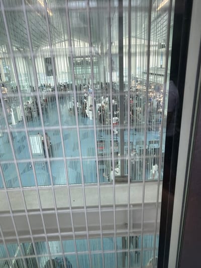 The image shows a view through a grate of a large, open indoor space, possibly an airport terminal or a convention center, in Ota, Japan. The space has a high ceiling with a complex architectural structure composed of reflective metal beams and panels, suggesting a modern design. A light blue floor is visible, patterned with darker blue shapes. Numerous people are scattered throughout the space, many of whom appear to be sitting at tables, suggesting a seating or dining area. Some individuals are standing and walking. The lighting appears bright and even, indicating an indoor environment illuminated by artificial lights or ample natural light filtered through the ceiling structure. The perspective is from a slightly elevated position, looking down and across the open area.