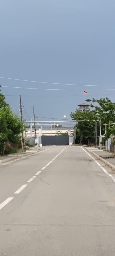 The image shows a view from a road leading towards a large gate. The road has white dashed lines marking the lanes. On the right side of the road, there's a curb and some broken pavement. Trees with green foliage line the left side of the road, and a wooden fence is visible behind them. In the background, there's a large, dark gray gate that appears to be closed. Above the gate, there are some industrial-looking structures and overhead wires. To the right of the gate, a tall tower stands with a flag at its top. The flag has red, blue, and orange stripes. The sky is overcast and gray, suggesting it might be late afternoon or early evening, or perhaps a cloudy day. There are no people visible in the image. The scene appears to be an entrance to a facility or compound, possibly industrial or military, given the gate and the tower with a flag. The location is identified as Vardanashen, Armenia.