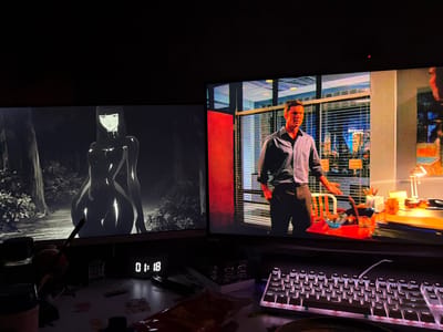 Two computer monitors are displayed in a dimly lit room, likely a home office or gaming setup. The monitor on the left shows a black and white animated character, a female with long dark hair and a form-fitting outfit, standing in a forest-like environment. The monitor on the right displays a live-action scene featuring a man in a striped shirt and dark pants, standing in an office setting with blinds and a city view through the window. In the foreground, a white keyboard with illuminated keys is visible, along with various items on a desk including a digital clock displaying "01:18", a pen, and snack packaging. The location in the city of Перепечино, Russia, is not discernible from the visual information provided in the image.