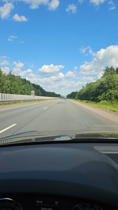 The image shows a view from inside a car driving on a highway. The road is asphalt with white lane markings and stretches into the distance, bordered by lush green trees and vegetation on both sides. The sky is a clear, bright blue with scattered white, fluffy clouds. The sun is shining, indicating it is daytime. On the right side of the frame, a portion of the car's dashboard is visible, including a digital display with the numbers "1414" and "07". There is also a dial that appears to be a speedometer, with markings up to "140". The overall scene evokes a sense of travel and open road. The location context provided is Уварово, Russia.