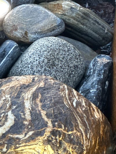 This media file is a close-up, overhead shot of a collection of smooth, rounded stones. The stones vary in size, shape, and color, with patterns that suggest natural geological processes.

The primary subjects are the stones themselves. Some are predominantly gray with lighter speckles, resembling granite. Others display striking bands of dark brown, black, and cream, creating a marbled or layered appearance. One large stone in the foreground has a prominent, swirling pattern of these colors. Another stone, elongated and appearing wet, shows distinct parallel lines or striations running along its length.

There is no activity or event taking place; the image is a still life composition of the stones.

Notable details include the varying textures and patterns of the stones, which are likely enhanced by being wet, as evidenced by the sheen on some surfaces. The lighting suggests it is daytime, possibly outdoors, given the natural subject matter. The setting appears to be a collection of stones, perhaps part of a decorative garden feature or natural landscape. No text is visible in the image. The location context is Givat Shmuel, Israel.