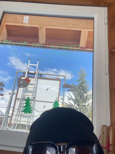 The image depicts a view from inside a structure, looking out through a white-framed window onto an alpine environment. In the immediate foreground, the top of an individual's head is visible, wearing a black knit beanie and large, dark-lensed sunglasses.
The window glass bears green marker inscriptions, reversed from this perspective, reading "= SMARTS 2 non Ramasse 2 = SMARTS 2 dans les CHAMAS". It is also decorated with multiple small black asterisk shapes, two green Christmas tree figures, two grey cloud shapes, and a black Hello Kitty head featuring a red bow.
Beyond the window, under a clear blue sky with scattered white clouds, a metallic ladder is observed leaning against a larger metal framework that includes a prominent white rectangular panel. To the right of this structure stands a tall, mature conifer tree. The distant background reveals snow-capped mountains, indicative of the setting in La Salle-les-Alpes, France, with visible patches of snow and faint outlines of buildings featuring red roofs.
The interior surrounding the window includes a wooden frame, exposed wooden beams above the window, and a light-colored wooden wall panel to the right, fitted with several wooden pegs from which red string or elastic is suspended. The scene is brightly lit by daylight.