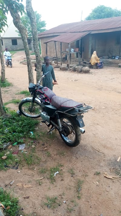 In Kagarko South, Nigeria, a dusty, unpaved road is the setting for this scene. In the foreground, a black motorcycle with a patterned seat is parked, leaning against a tree. To the left, a young boy stands near the motorcycle, facing the camera. Further in the background, a woman in a yellow headwrap and patterned skirt sits on a bench outside a modest dwelling with a corrugated metal roof. Another individual, presumably a man, is visible on a white motorcycle in the distance, riding away from the camera. The environment appears rural, with sparse vegetation and scattered debris on the ground. The buildings are simple, constructed with mud or concrete blocks.
