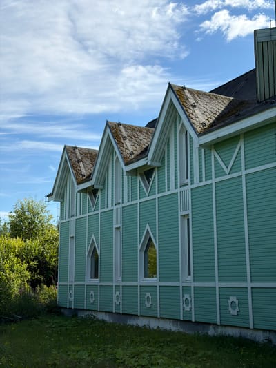 A light green, wooden building with white trim stands in an outdoor environment under a partly cloudy blue sky. The building has a multi-gabled roof covered in gray shingles that show patches of moss. Several narrow, white-framed windows are visible on the building's facade, some with pointed arch tops. The siding of the building is horizontal and painted a light green color, with white decorative elements and trim. To the left of the building, there is lush green vegetation and trees. The foreground consists of a grassy area with some smaller plants and flowers. The image appears to be taken from a slightly low angle, looking up at the building. The building is located in the city of Läskelä, Russia.