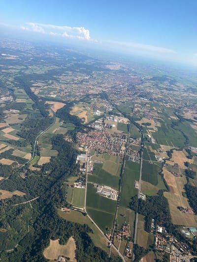 This is an aerial photograph of a rural landscape in Pombia, Italy, likely taken from an airplane. The image shows a patchwork of agricultural fields, forests, and a small town with red-tiled roofs. A canal or river runs through the center of the frame, bordered by trees. In the distance, a hazy horizon and a clear blue sky suggest fair weather. The overall scene depicts a tranquil, agrarian environment.