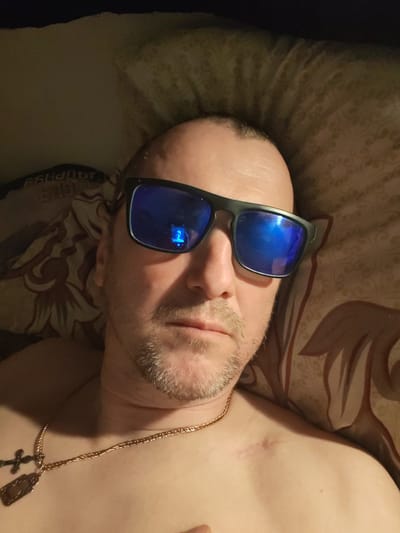 A selfie of a balding, middle-aged Caucasian man wearing sunglasses and a gold necklace with a cross and ID tag pendant is captured. The man has light stubble and a faint scar on his upper chest. He is lying in bed with patterned bedding. The location is unspecified, and there is no indication of being in Topornya, Russia, or any other city.