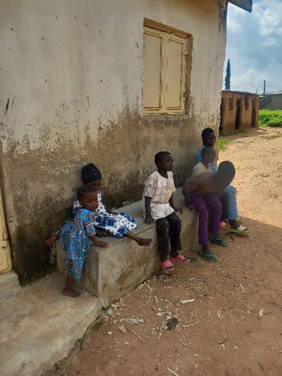 The scene is located in Zangon Aya, Nigeria, depicting six children positioned in front of a weathered building under a partly cloudy sky. The single-story structure features an off-white or light-colored plastered facade, exhibiting significant peeling paint and grime, particularly on the lower half. A closed, aged yellow wooden window with two vertical panes is present on the wall. A concrete bench extends along the building's base. The immediate foreground consists of reddish-brown dirt, sparsely covered with organic debris and small fragments.
Six children are present. From left to right:
1. A toddler, barefoot and wearing a blue and white patterned full-body garment, stands on a lower concrete step, leaning forward with hands placed on the concrete bench.
2. An individual, likely a female child, wearing a white dress with blue patterns and a dark head covering, sits on the concrete bench.
3. A boy, wearing a light-colored long-sleeved shirt, dark pants, and pink open-toed sandals, sits on the concrete bench.
4. A boy, wearing a light blue short-sleeved shirt and blue pants, with light green flip-flops, sits behind the previous child on the concrete bench.
5. A boy, wearing a light brown short-sleeved shirt and purple pants, with green flip-flops, sits on the concrete bench.
6. A partially visible boy, wearing light-colored clothing and light green flip-flops, sits on the dirt ground at the far right.
All children are generally oriented towards the right side of the frame. In the background, several rudimentary structures constructed from unmortared mud or clay bricks are visible, along with a tall, slender green tree and a utility pole against the sky.