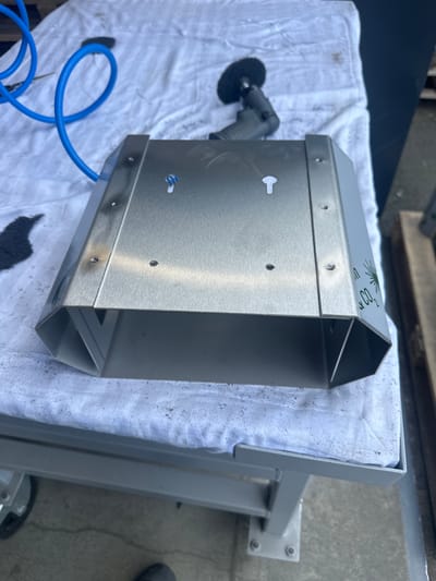 This is a medium-angle, close-up shot of a metal object, likely a ventilation component, resting on a white cloth spread over a grey metal workbench. The object is trapezoidal in shape with an open bottom. It appears to be made of brushed stainless steel, with visible rivets and screw holes along its sides. Some scorch marks are visible on the top surface.
To the left of the object, a bright blue coiled pneumatic hose is visible, attached to an unseen air compressor. Above the object, a black air-powered grinding tool with a circular wire brush attachment is positioned on the cloth.
On the right side of the object, green lettering is visible, partially obscured. The visible portion appears to read "CO2".
The scene is outdoors, with a concrete floor visible beneath the workbench. The background is blurred, but suggests an industrial or workshop setting, possibly a loading dock or outdoor storage area. There are no people visible in the image. The overall impression is of a work-in-progress or recently completed fabrication, with tools and materials laid out.
The location is identified as Loo, Estonia.